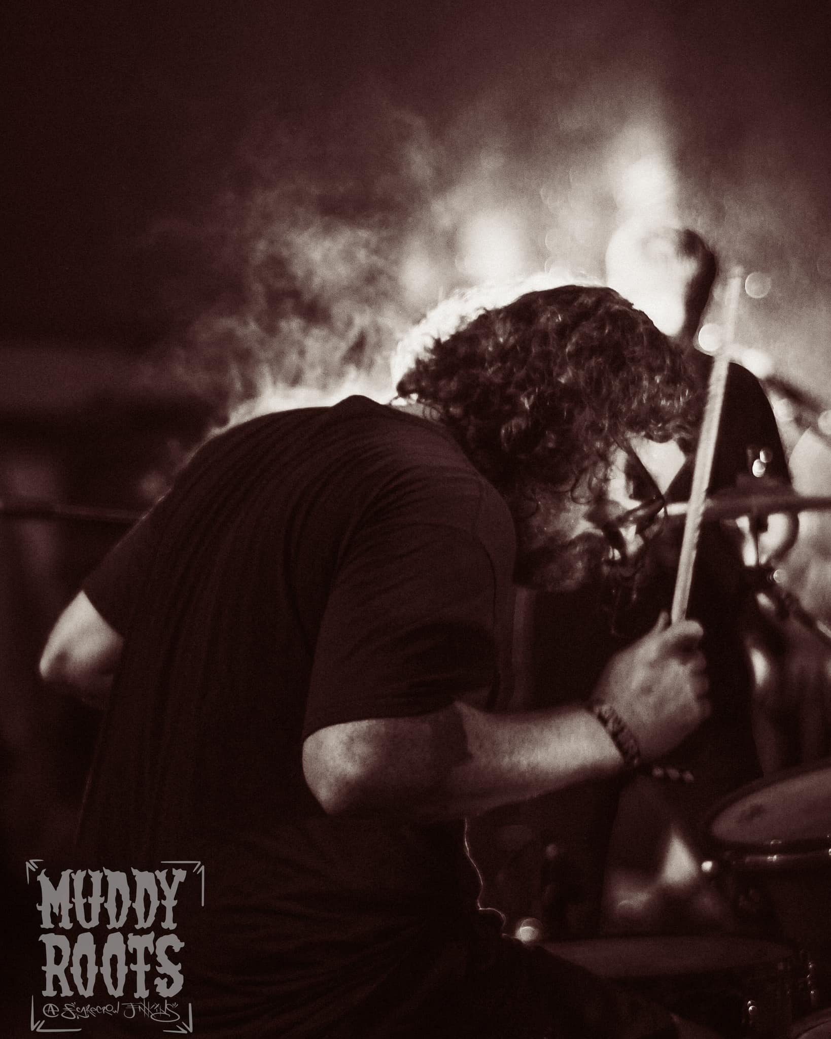 A stylized greyscale photo of Daniel playing drums at the Muddy Roots festival in 2024